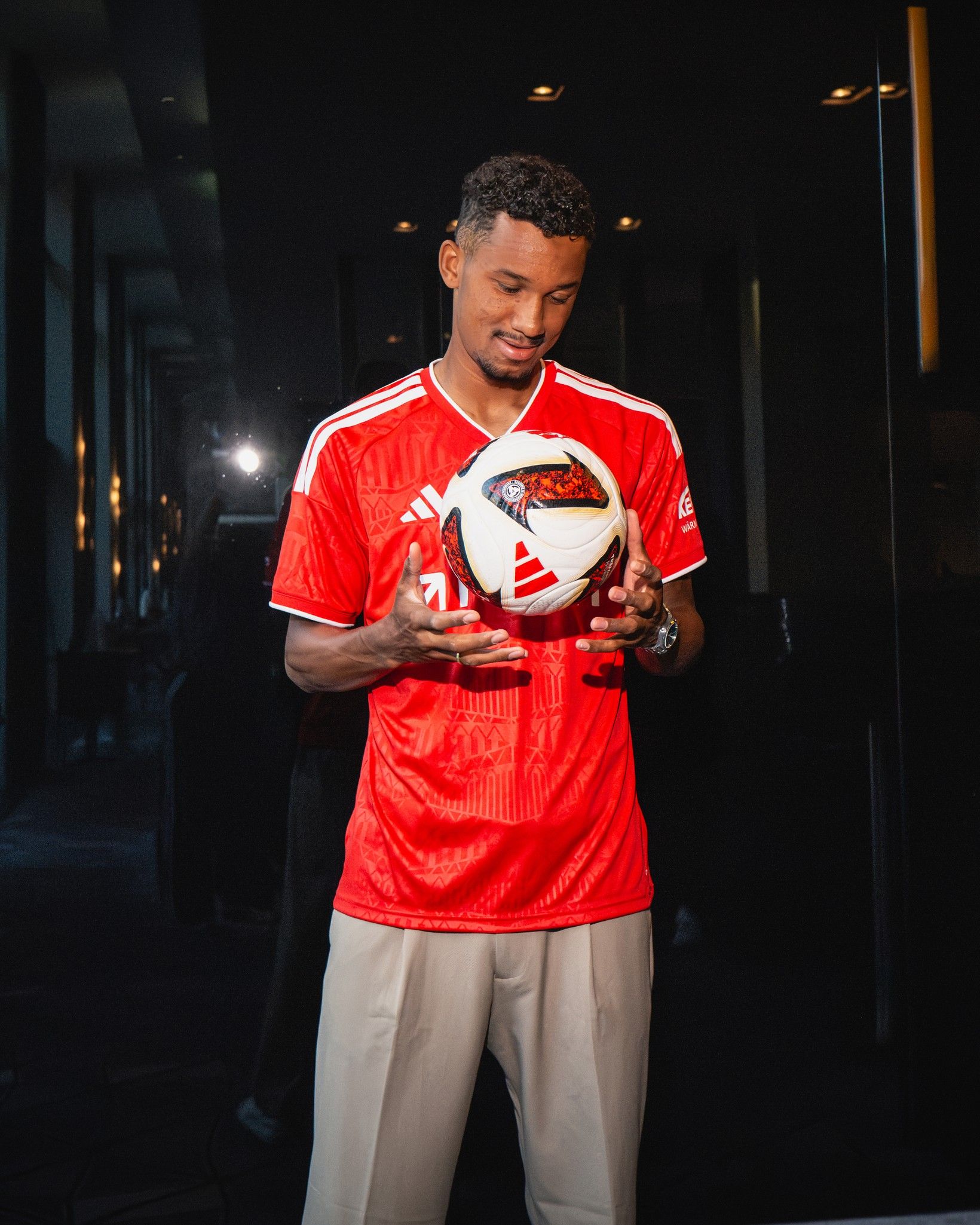 Ein junger Mann in einem roten Fußballtrikot hält einen Fußball in den Händen und schaut fasziniert darauf.