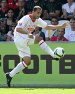 Ein Fußballspieler in weißem Trikot schießt den Ball während eines Spiels. Zuschauer im Hintergrund sind zu sehen.