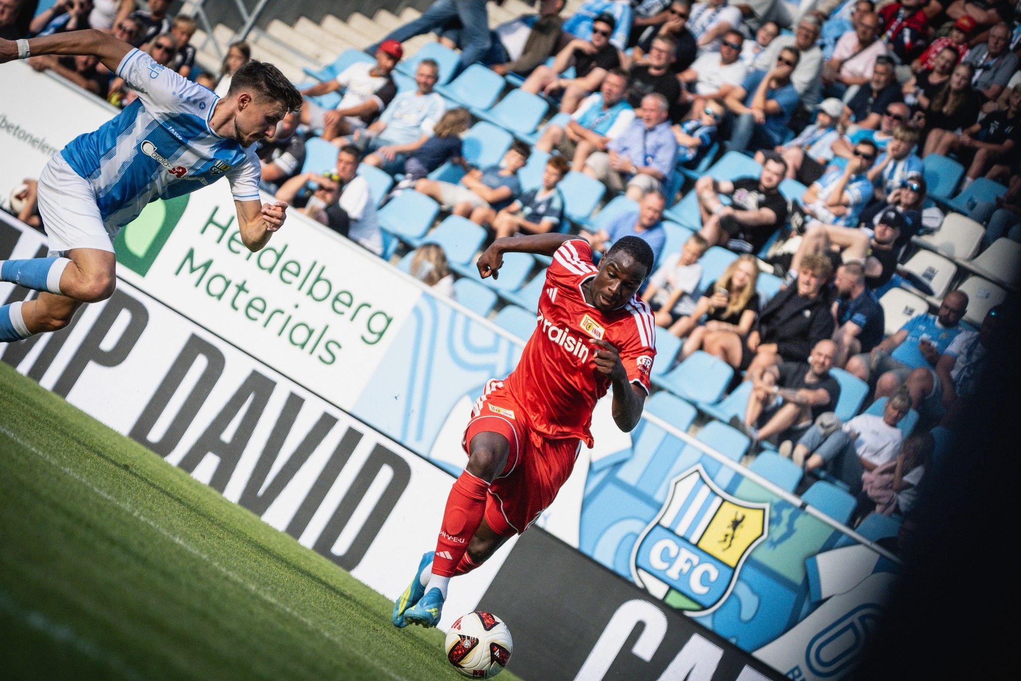 Ein Fußballspieler in roter Kleidung dribbelt den Ball, während ein blauer Spieler ihn verfolgt. Zuschauer sitzen im Hintergrund.
