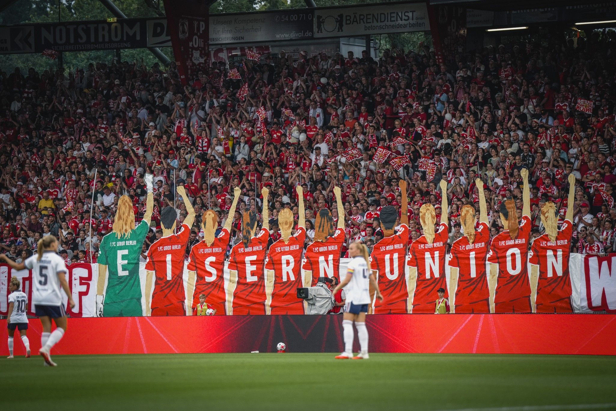 Fankurve mit Banner der Mannschaft "Eisen Union" und jubelnden Fans in roter Kleidung. Im Vordergrund Spielerinnen auf dem Spielfeld.