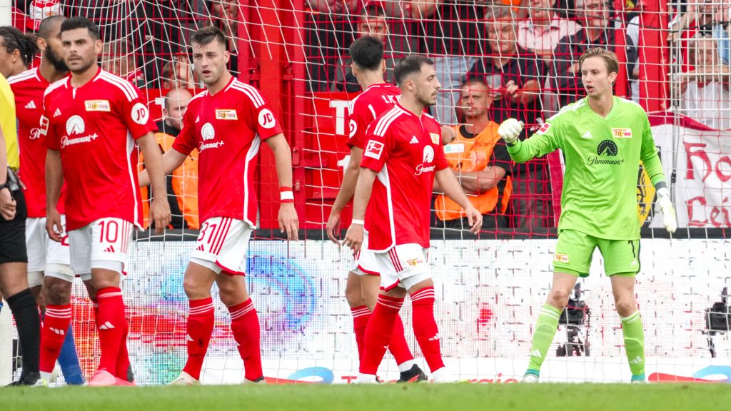 Soccer team in red jerseys on the field, with goalkeeper in green gear and players in action.