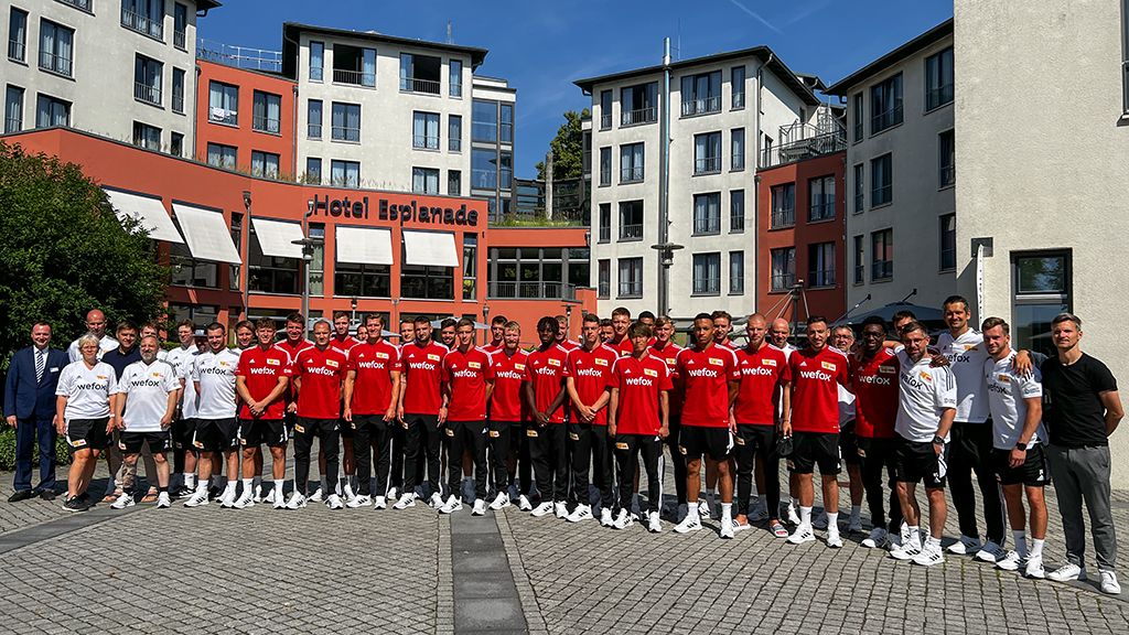 A group of soccer players in red jerseys stands in front of a hotel, surrounded by several buildings.