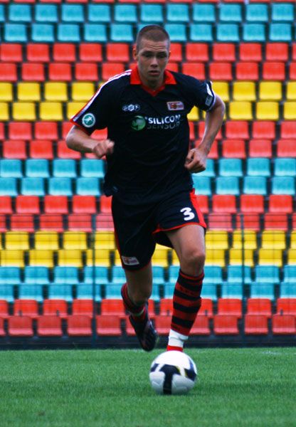 Ein junger Fußballspieler in schwarzem Trikot läuft mit dem Ball auf einem farbenfrohen Stadionrand.