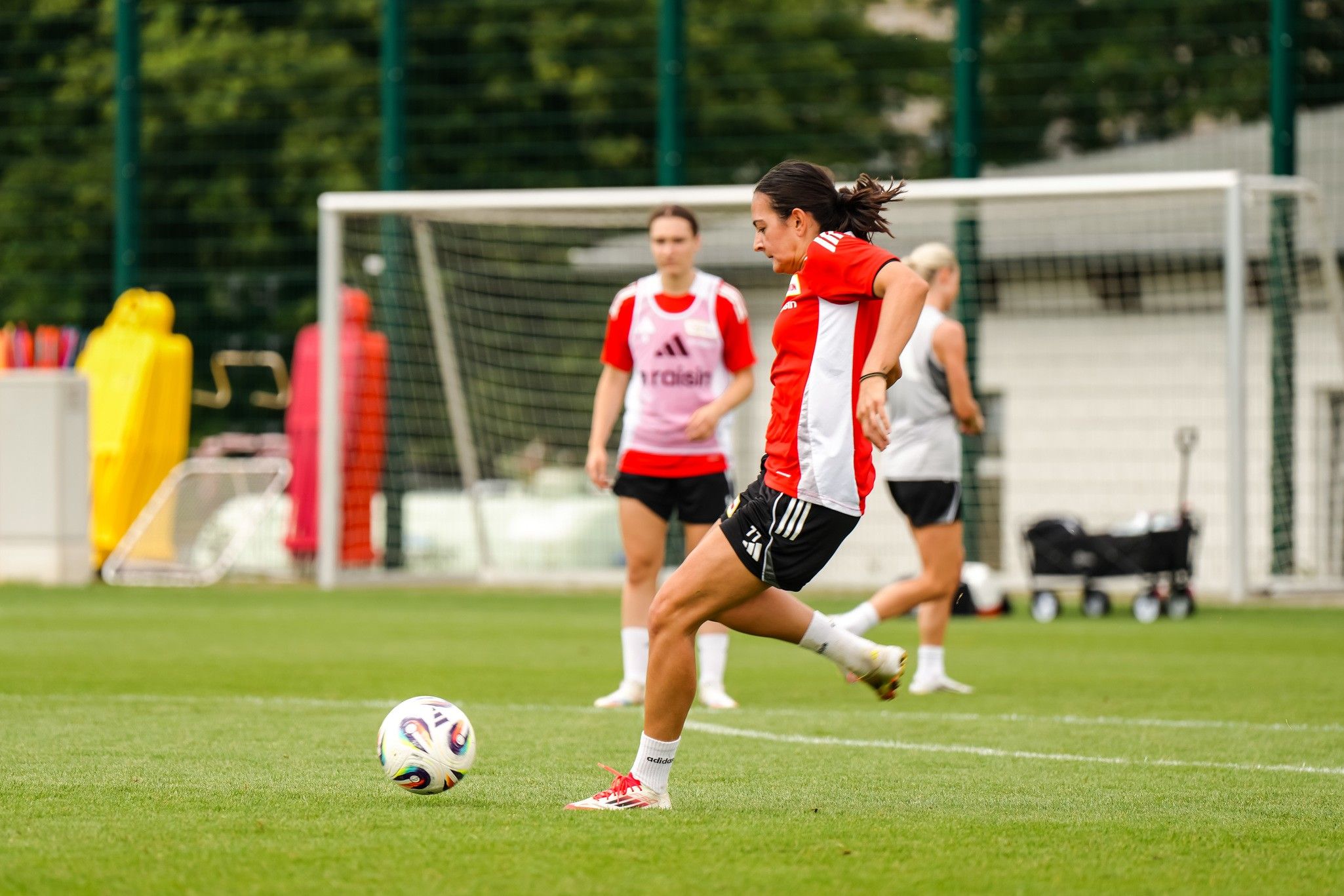 Eine Fußballspielerin in rotem Trikot tritt den Ball während des Trainings, im Hintergrund weitere Spielerinnen.