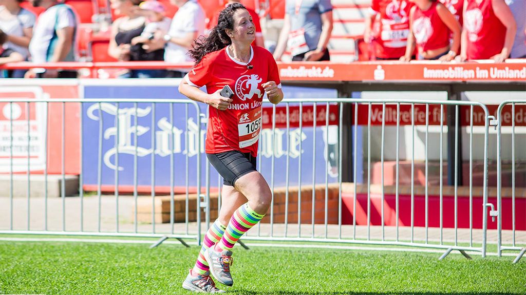 A woman in a red-and-white sports outfit happily runs on a grassy area in front of spectators, wearing colorful striped socks.