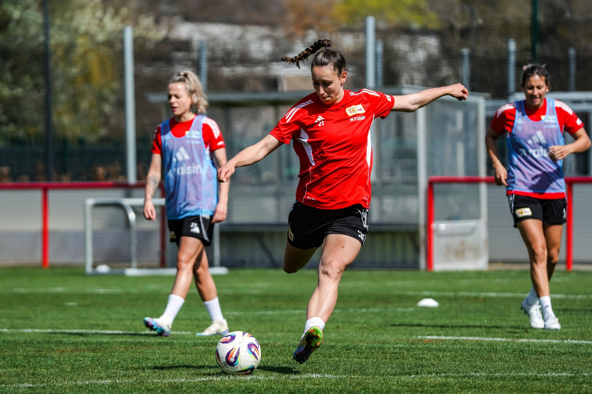 Drei Frauen in roten Trikots trainieren auf einem Fußballplatz. Eine Spielerin schießt den Ball.