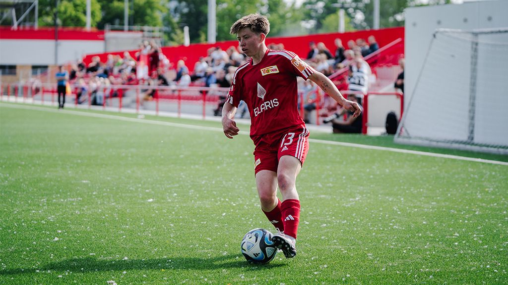 Ein Fußballspieler in roter Uniform dribbelt den Ball auf einem Rasenplatz, während Zuschauer im Hintergrund zuschauen.