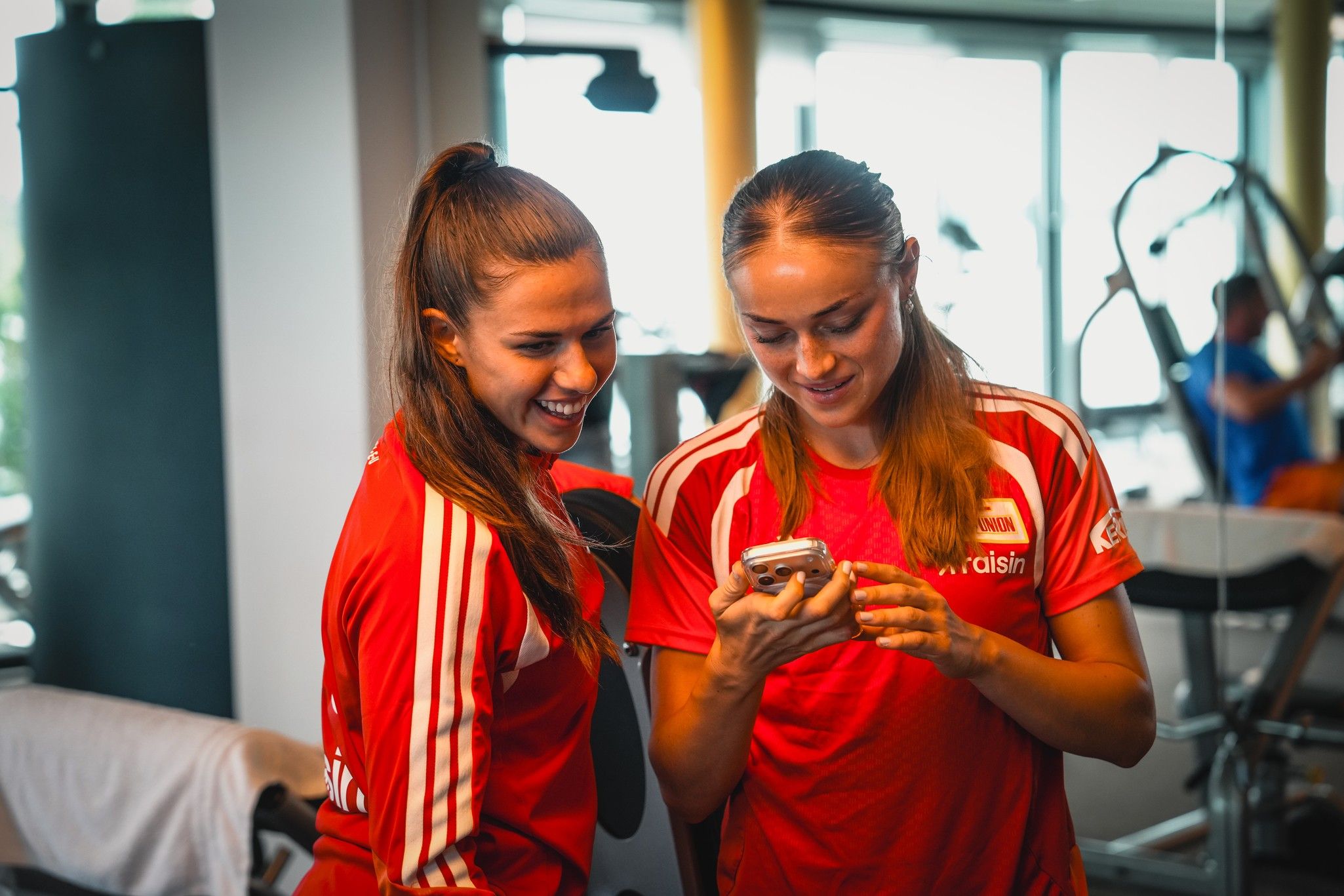 Zwei Frauen in roten Sporttrikots schauen auf ein Smartphone und lächeln, während im Hintergrund ein Fitnessstudio zu sehen ist.