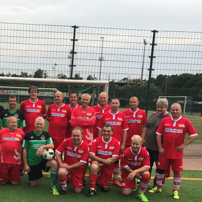 Eine Gruppe von 16 Fußballspielern in roten Trikots steht auf einem Fußballplatz vor dem Tor.