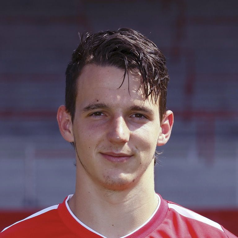 Ein junger Mann mit dunklem, lockigem Haar trägt ein rotes Sporttrikot und lächelt in die Kamera.