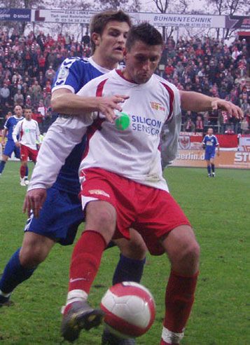 Zwei Fußballspieler kämpfen um den Ball während eines Spiels auf einem grünen Rasenfeld, im Hintergrund eine jubelnde Menge.