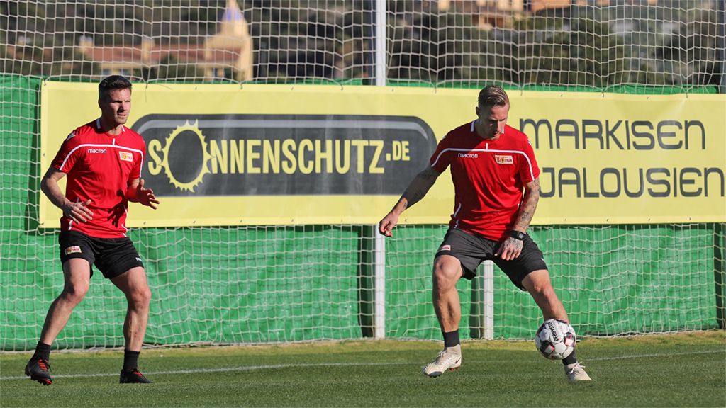 Zwei Fußballspieler in roten Trikots trainieren mit einem Ball auf einem Sportplatz, vor einem Werbebanner.