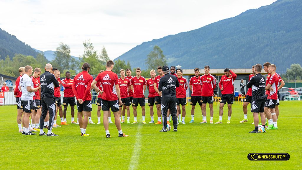 Gruppe von Fußballspielern in roten Trikots versammelt sich zu einer Besprechung auf einem Trainingsplatz, Berge im Hintergrund.