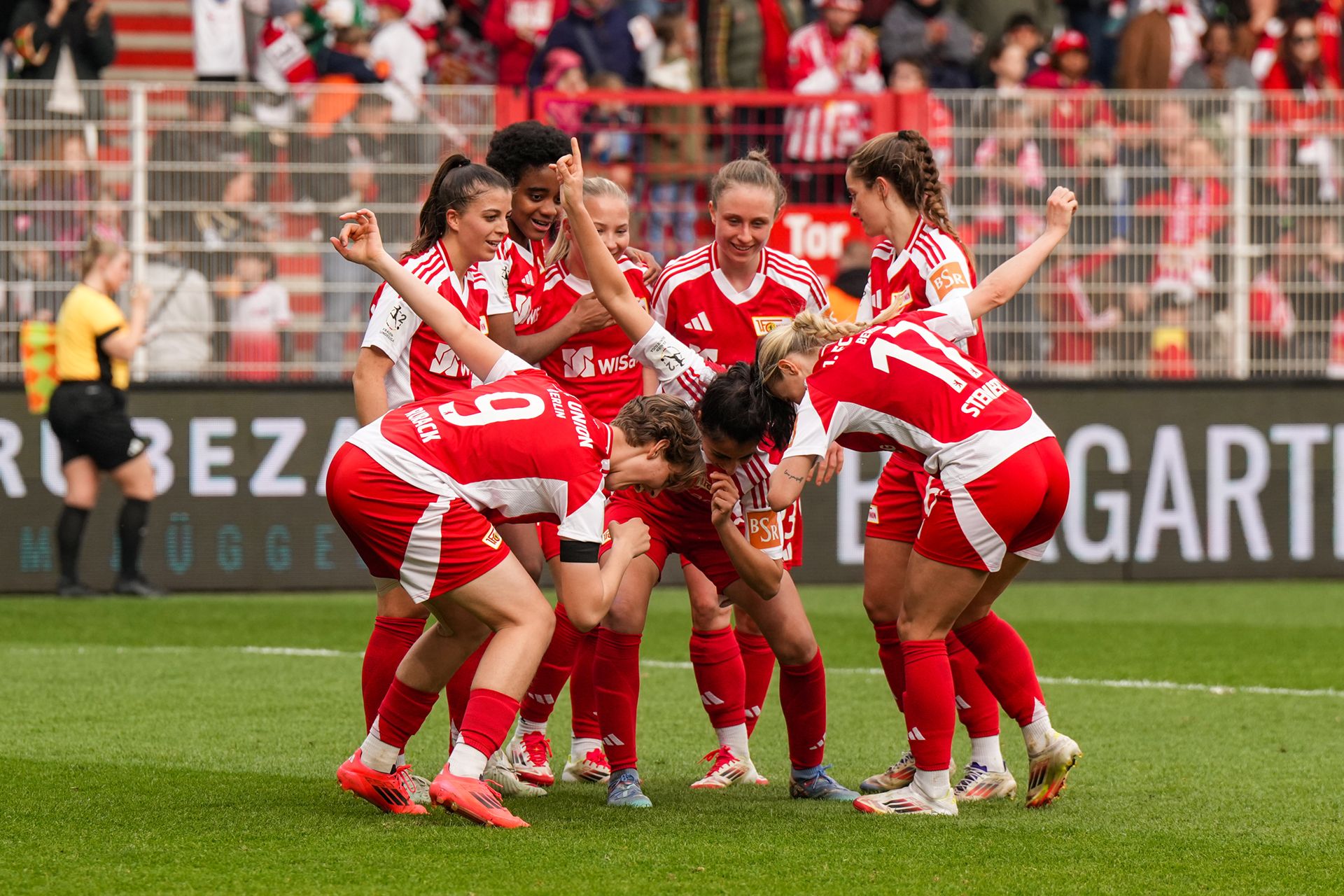 Gruppenkuscheln von Spielerinnen in roten Fußballtrikots nach einem Tor, auf einem Rasenplatz mit Zuschauern im Hintergrund.
