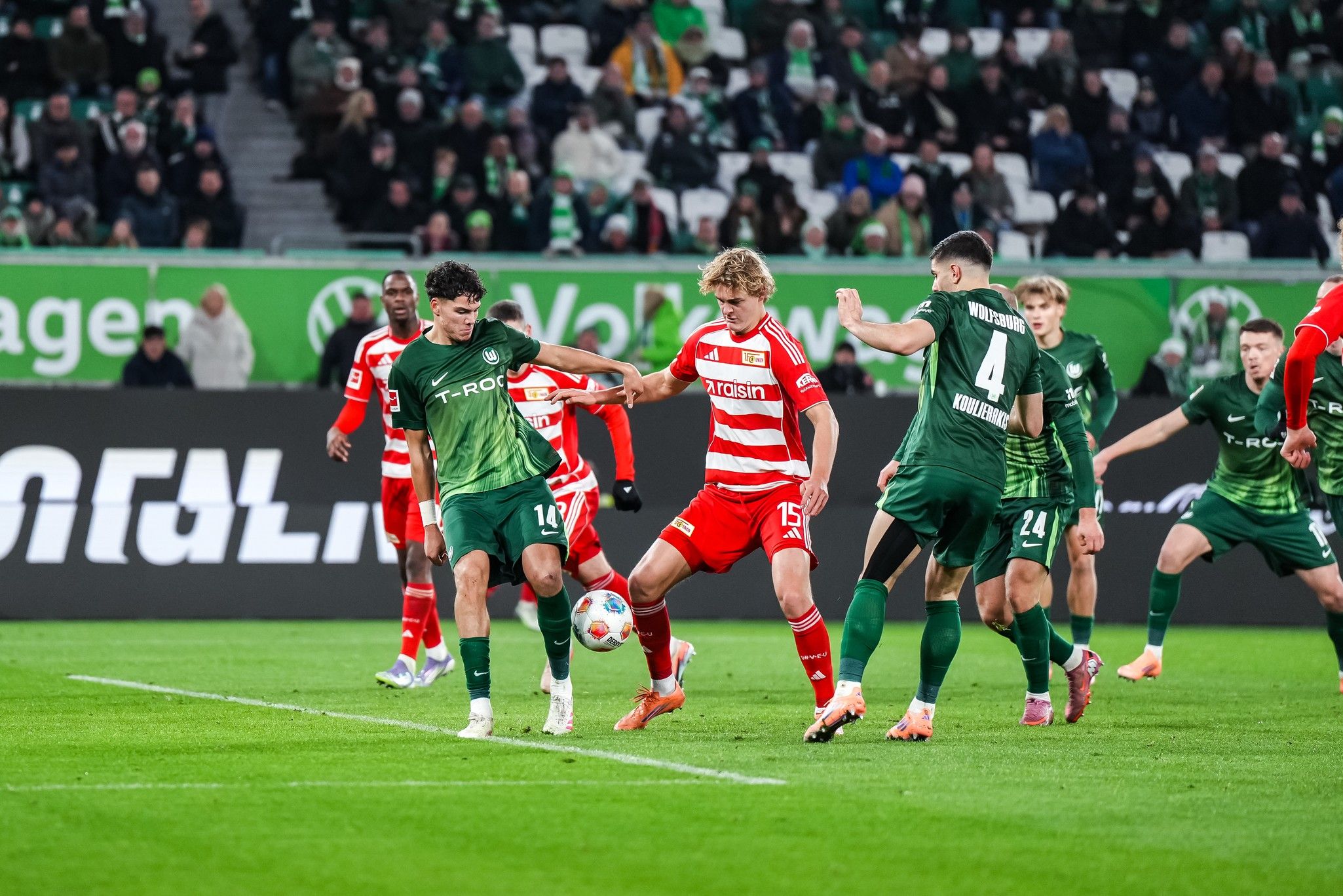 Zwei Fußballmannschaften kämpfen um den Ball während eines Spiels in einem Stadion mit jubelnden Fans im Hintergrund.