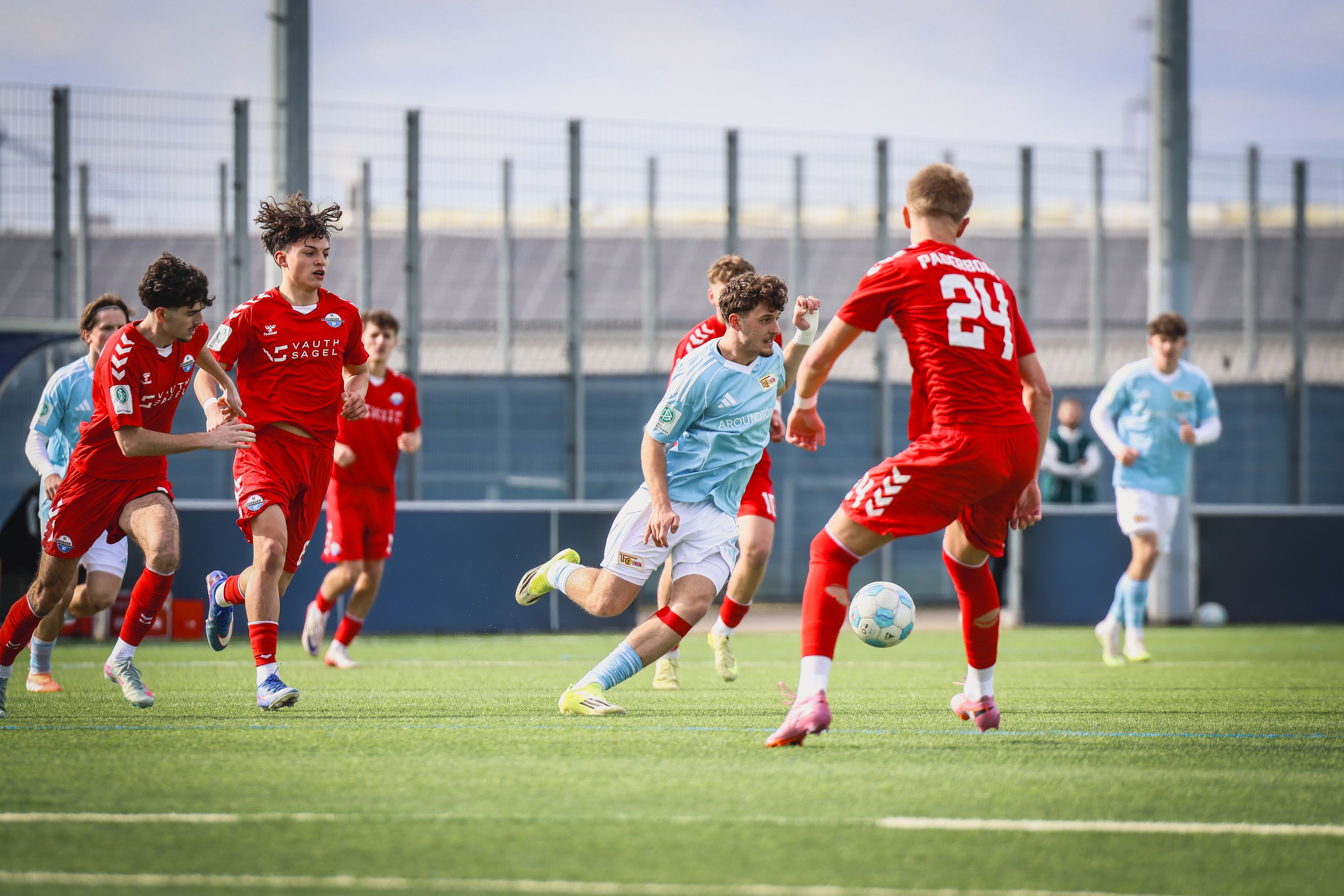 Zwei Fußballmannschaften im Wettkampf auf einem grünen Kunstrasenplatz. Spieler in blauen und roten Trikots sind aktiv am Ball.