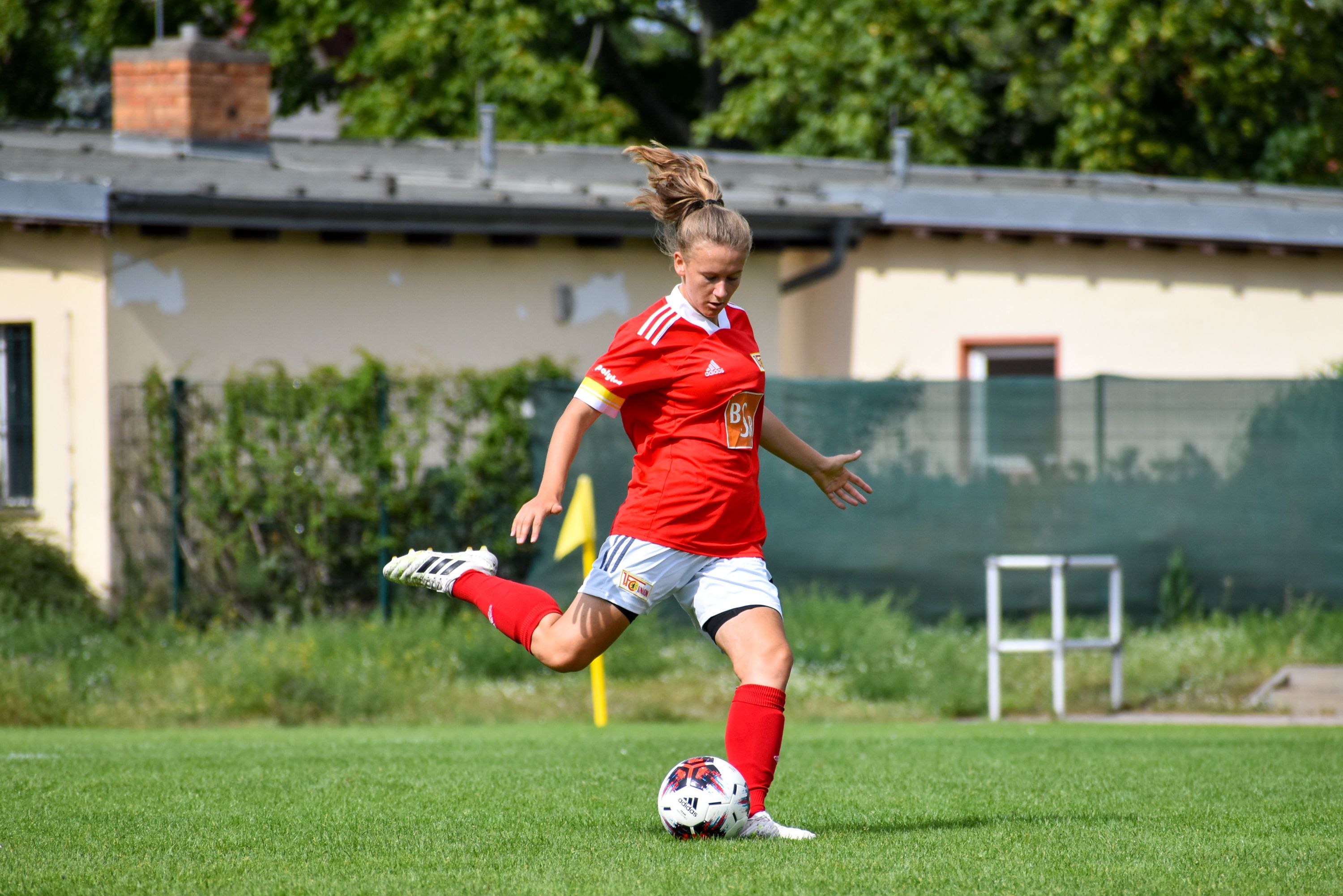 Eine Fußballspielerin in rotem Trikot schießt den Ball auf einem grünen Rasenplatz. Im Hintergrund sind Gebäude und Bäume.