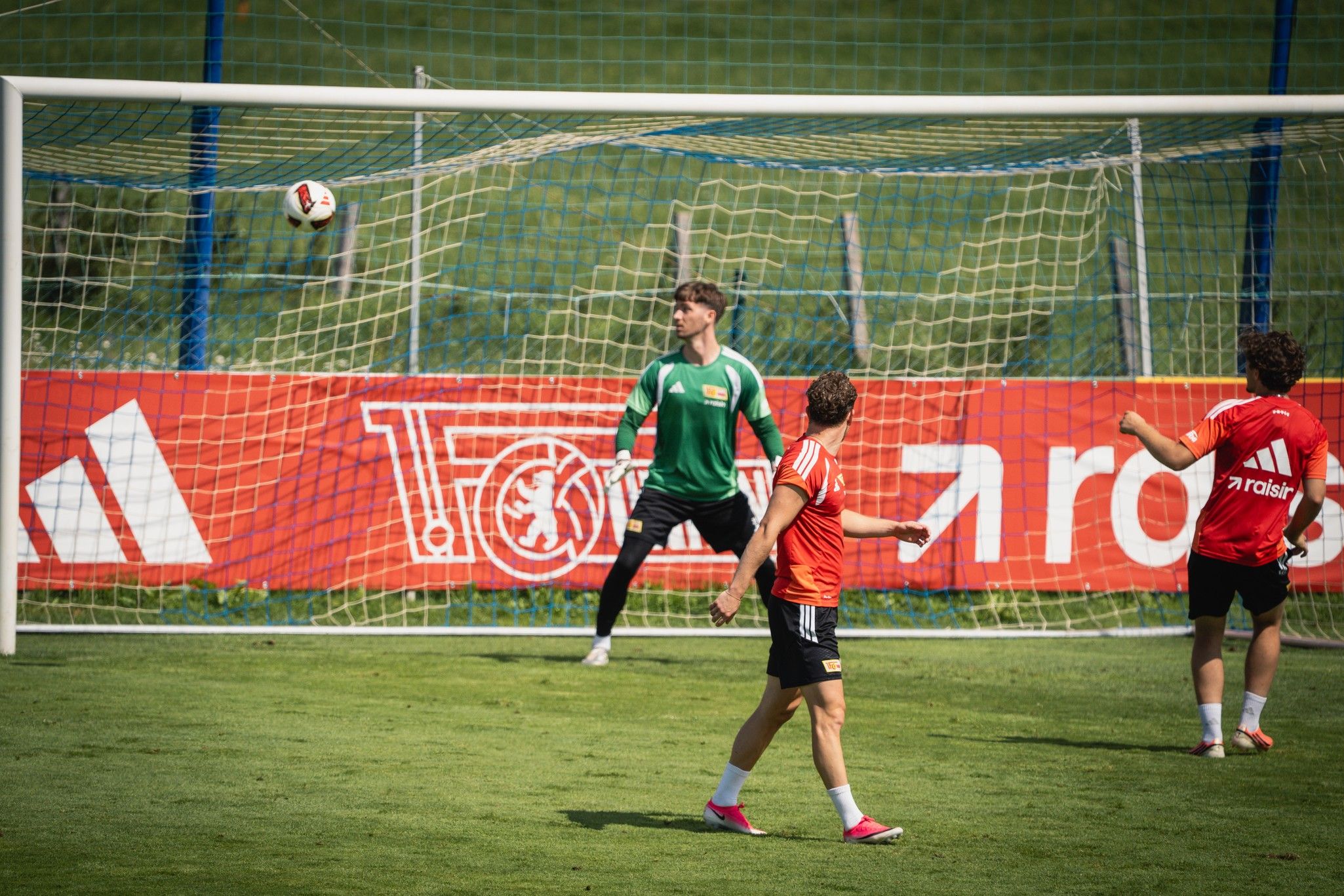 Ein Fußballtor mit einem Torwart in grün, der einen Ball abwehrt; zwei Spieler in roten Trikots stehen in der Nähe.