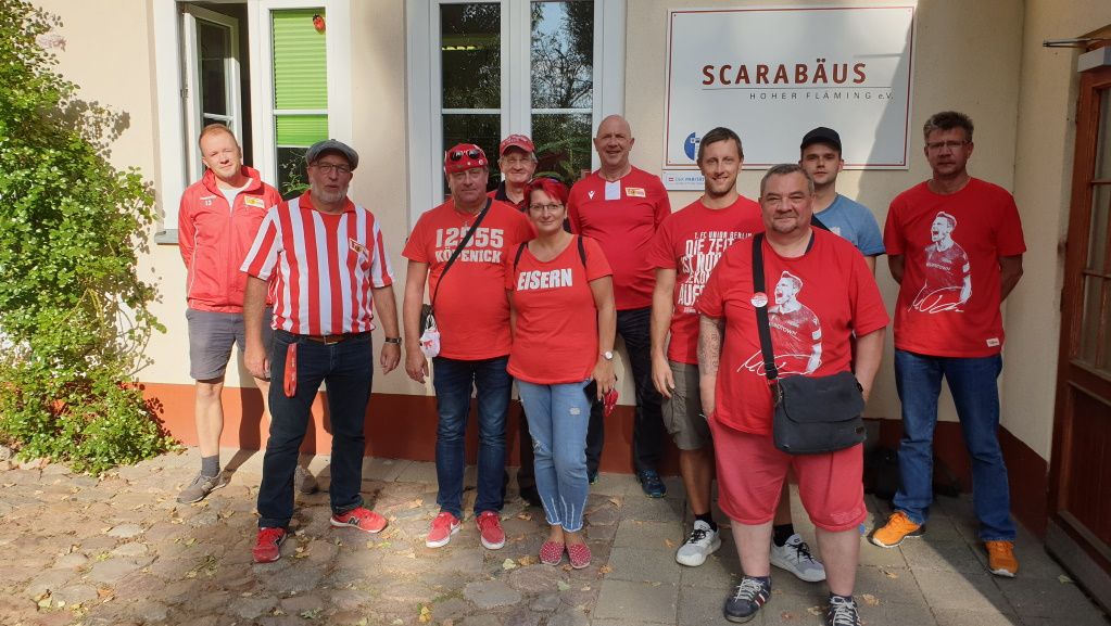 Gruppe von Menschen in roten T-Shirts vor einem Gebäude mit dem Schriftzug "SCARABÄUS", lächelnd und stehend.