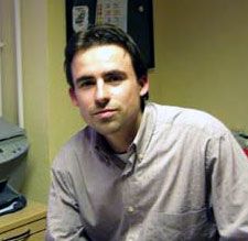 Ein Mann mit dunklen Haaren sitzt in einem Büro, trägt ein helles Hemd und schaut direkt zur Kamera.