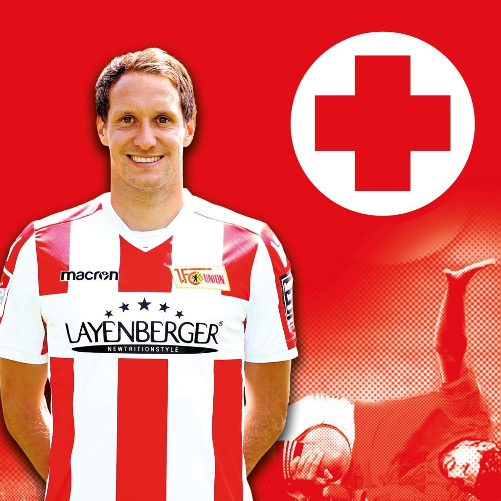 Lächelnder Fußballspieler in rot-weißem Trikot vor rotem Hintergrund mit einem Kreuz-Symbol.