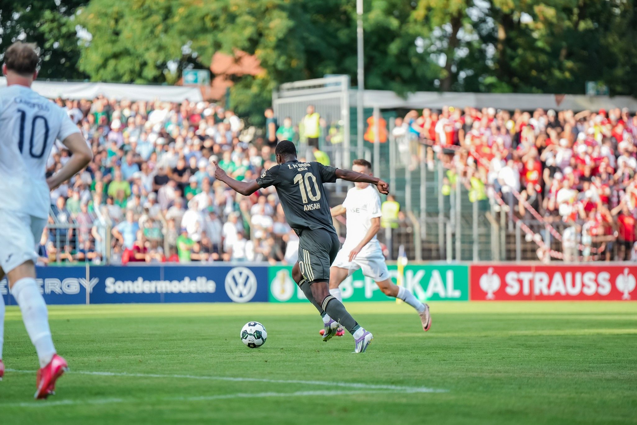 Ein Spieler in schwarz mit der Nummer 10 dribbelt den Ball auf einem Fußballfeld, während Zuschauer im Hintergrund zuschauen.