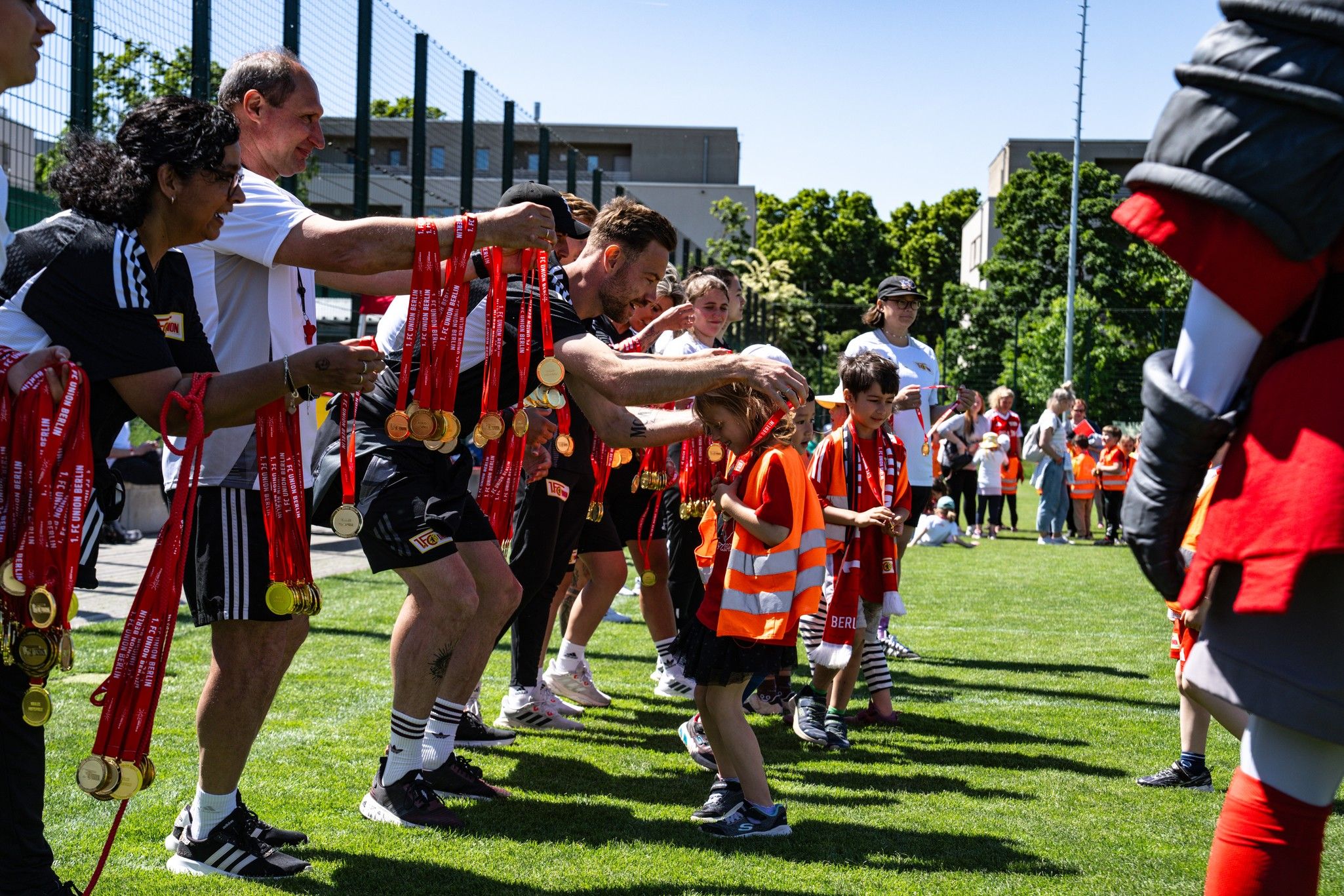 Sportveranstaltung mit Kindern, die Medaillen von Trainern und Betreuern erhalten. Teilnehmer tragen orangene Westen und Medaillen.