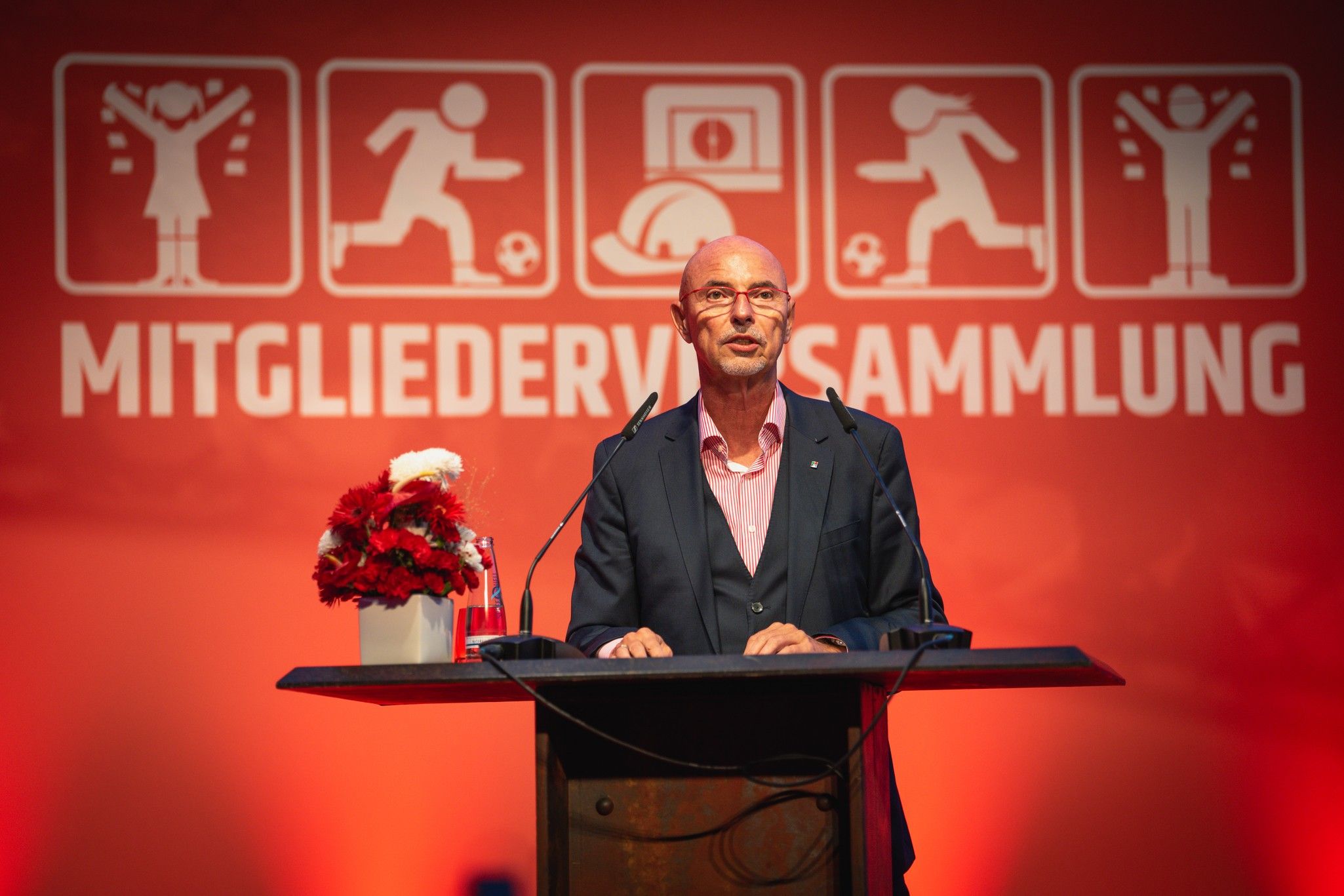 Mann steht am Rednerpult während einer Veranstaltung mit rotem Hintergrund und Symbolen von sportlichen Aktivitäten.