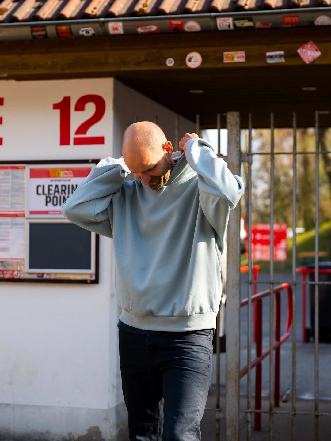 Mann mit Glatze trägt einen hellblauen Pullover und steht an einem Eingangsbereich mit einem Schild und einem Gitter.