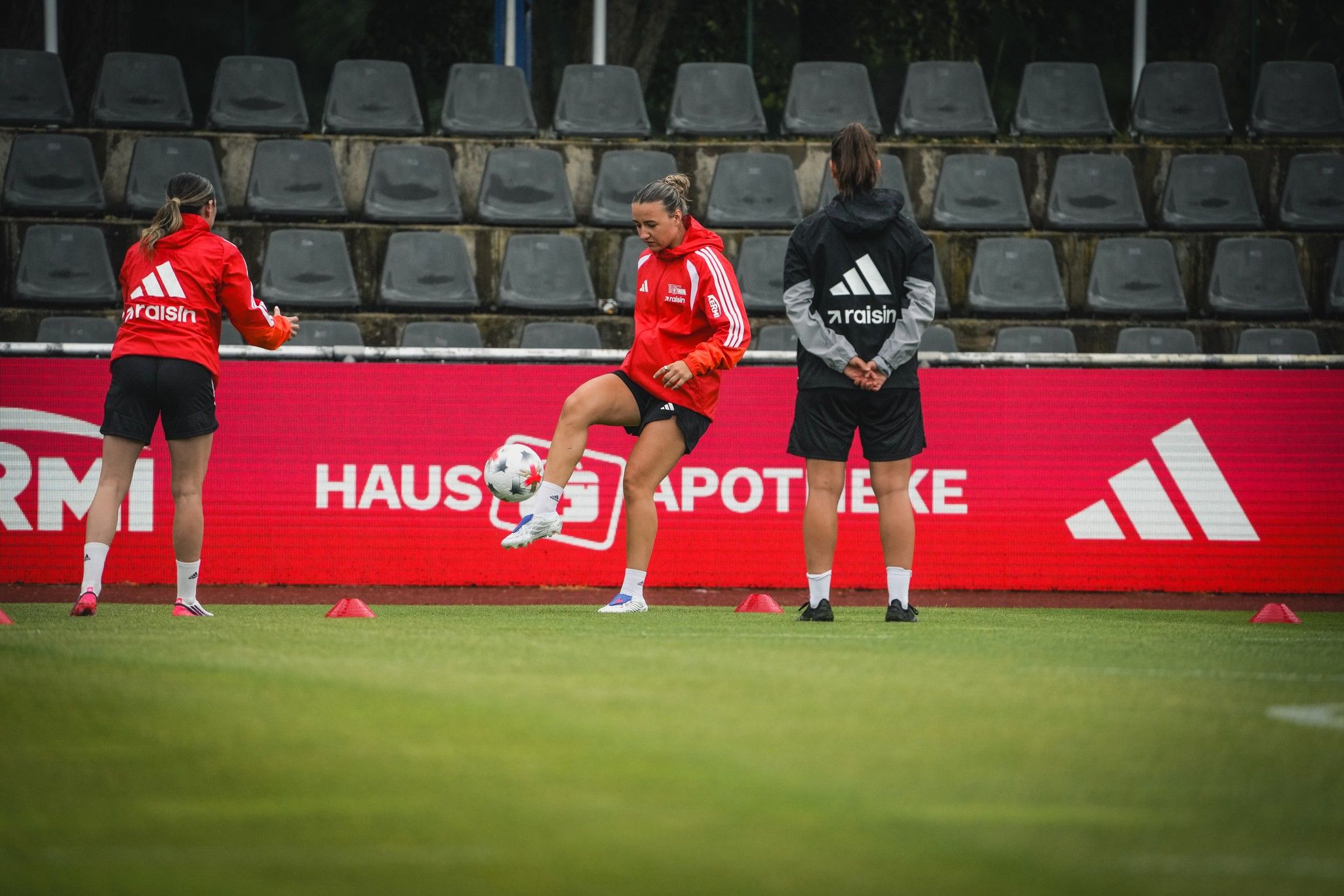 Drei Fußballspielerinnen in roten Trikots trainieren auf dem Platz, während sie Pylonen umrunden und den Ball jonglieren.