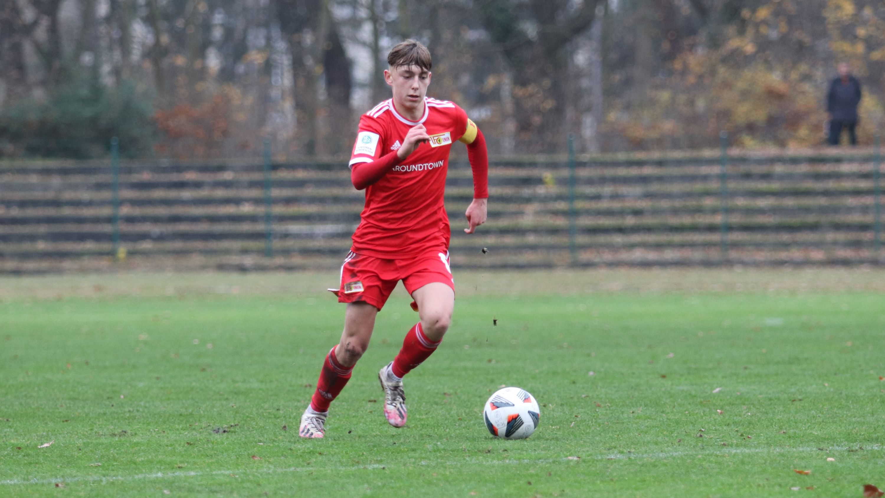 Ein junger Fußballspieler in rotem Trikot sprintet mit dem Ball auf einem grünen Spielfeld.