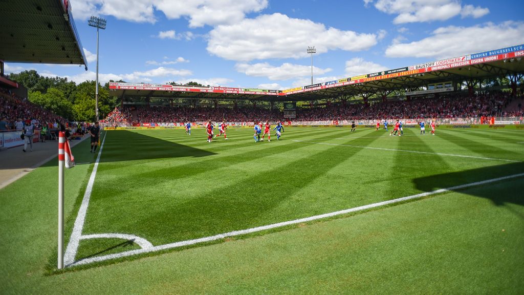 Football match in a stadium, spectators in the stands, well-maintained grass, and clouds in the sky.