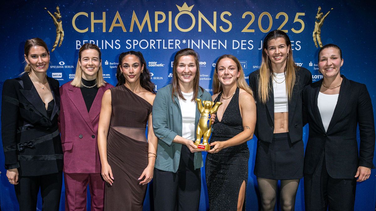 Gruppe von sieben Sportlerinnen, die stolz eine Trophäe halten, vor einer blauen Wand mit der Aufschrift "Champions 2025".