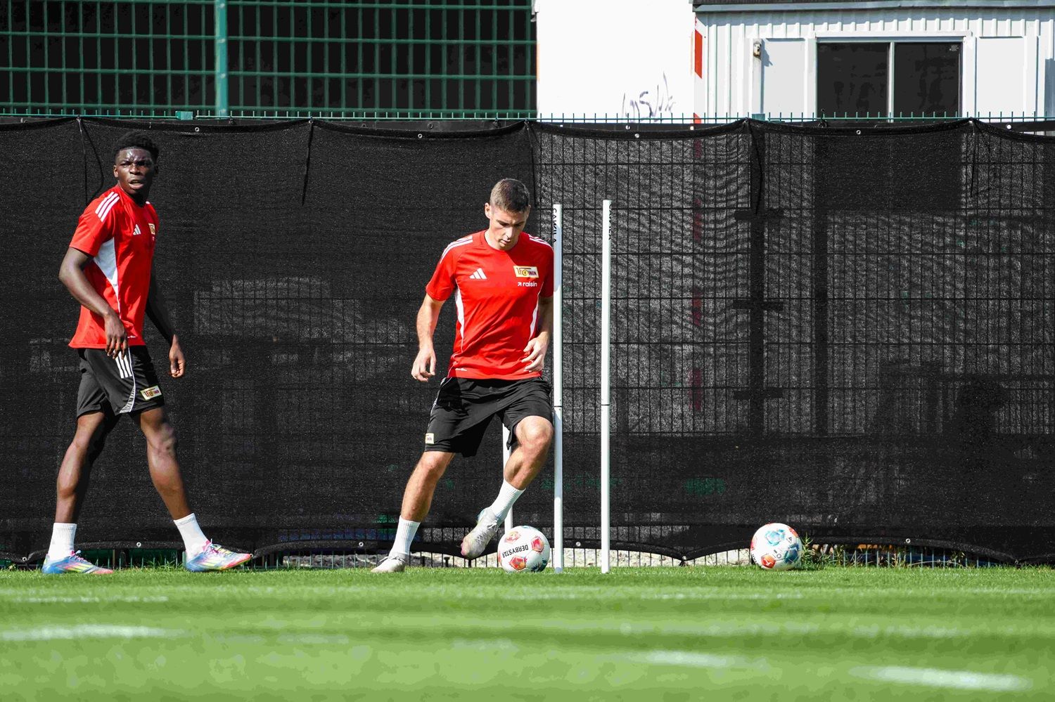 Zwei Fußballspieler in roten Trikots trainieren auf einem Rasenplatz, umgeben von schwarzen Sichtschutznetzen.
