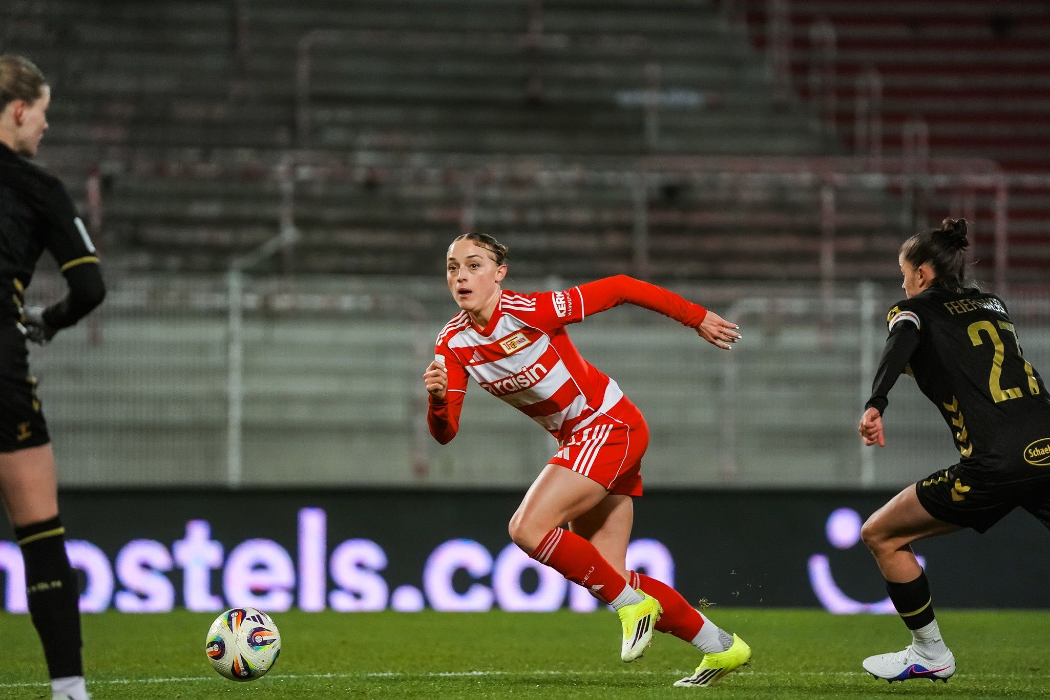 Eine Fußballspielerin in rot weicht der gegnerischen Verteidigung aus und läuft mit dem Ball in Richtung Tor.