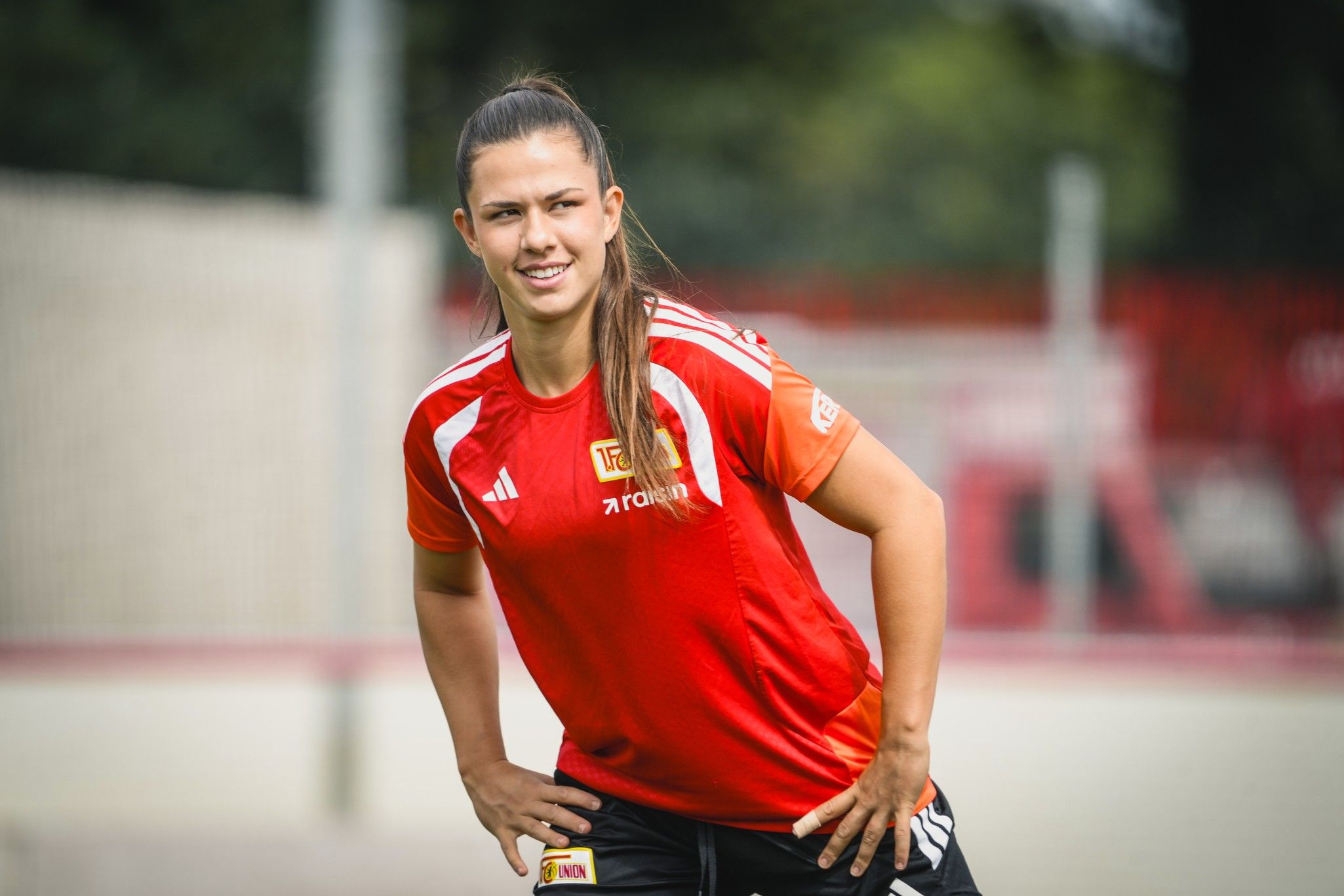 Eine sportliche Frau in rotem Trikot steht mit entspannter Pose auf einem Trainingsgelände.