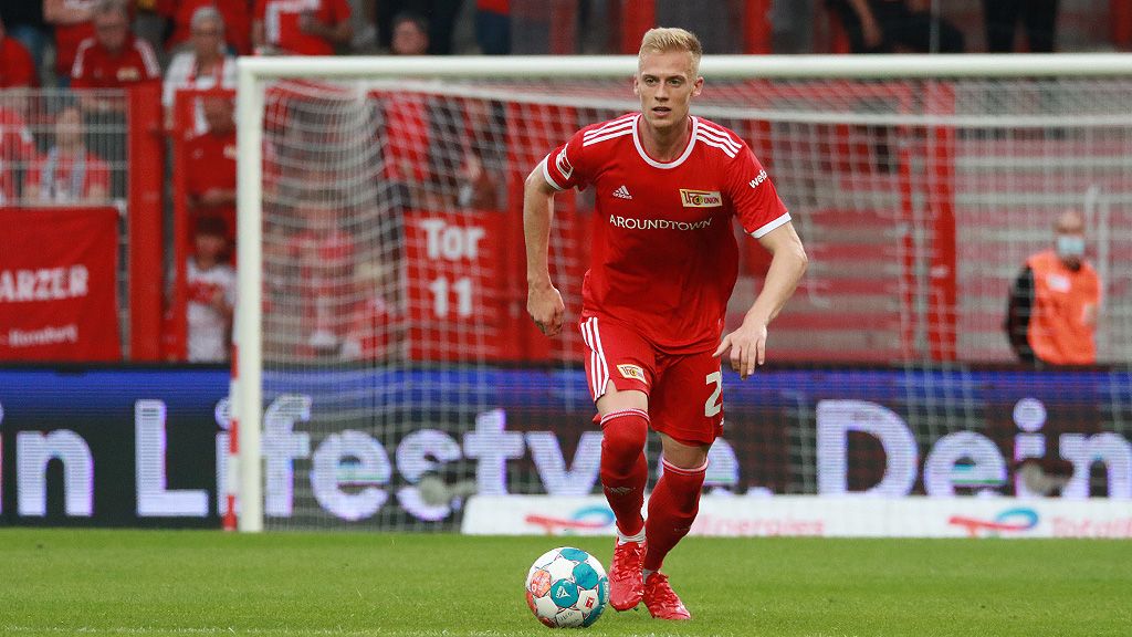 A soccer player in a red jersey dribbles the ball during a game on the field. Fans can be seen in the background.
