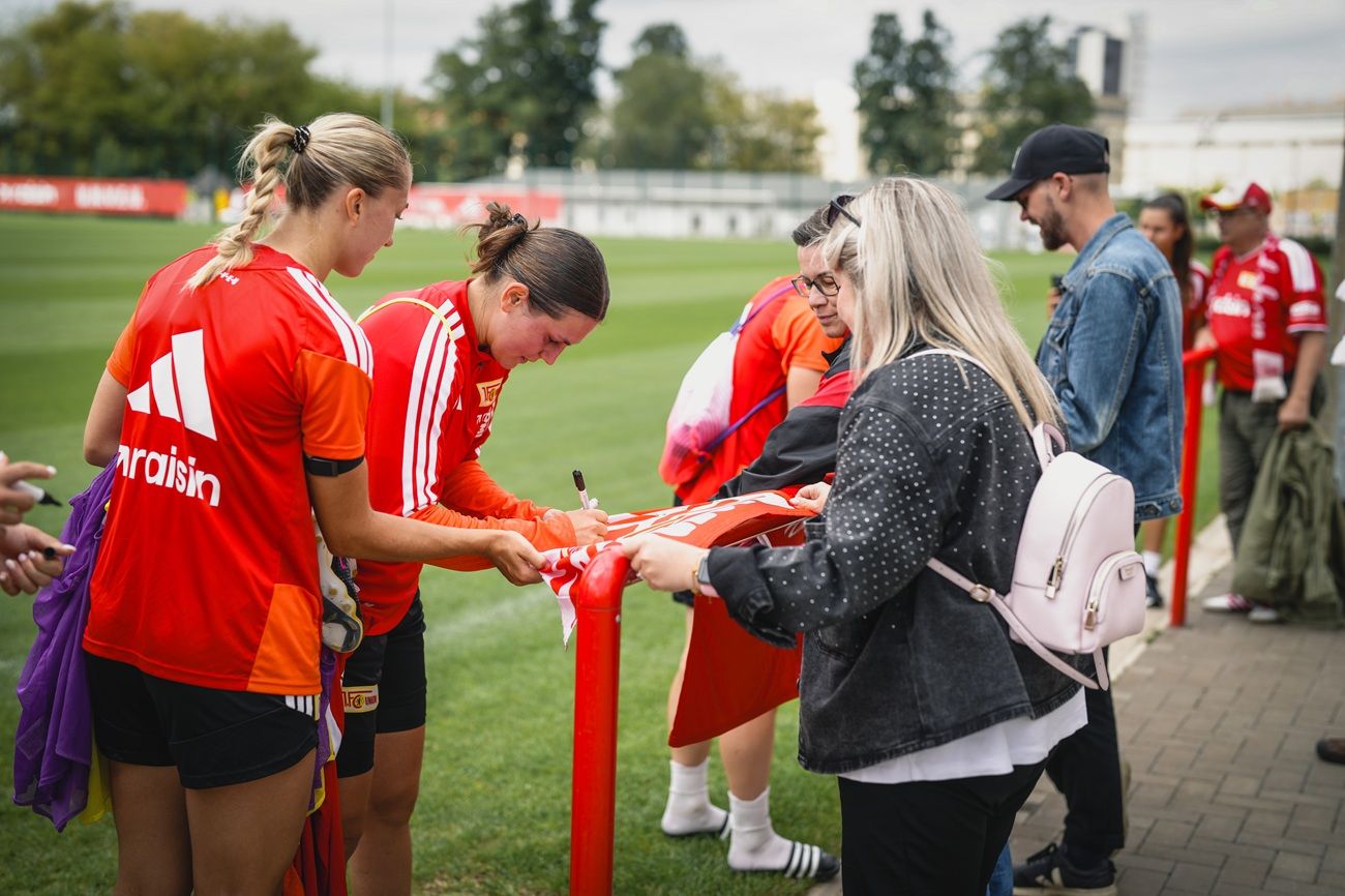 Zwei Fußballspielerinnen stehen auf einem Trainingsplatz und signieren ein Trikot für Fans.