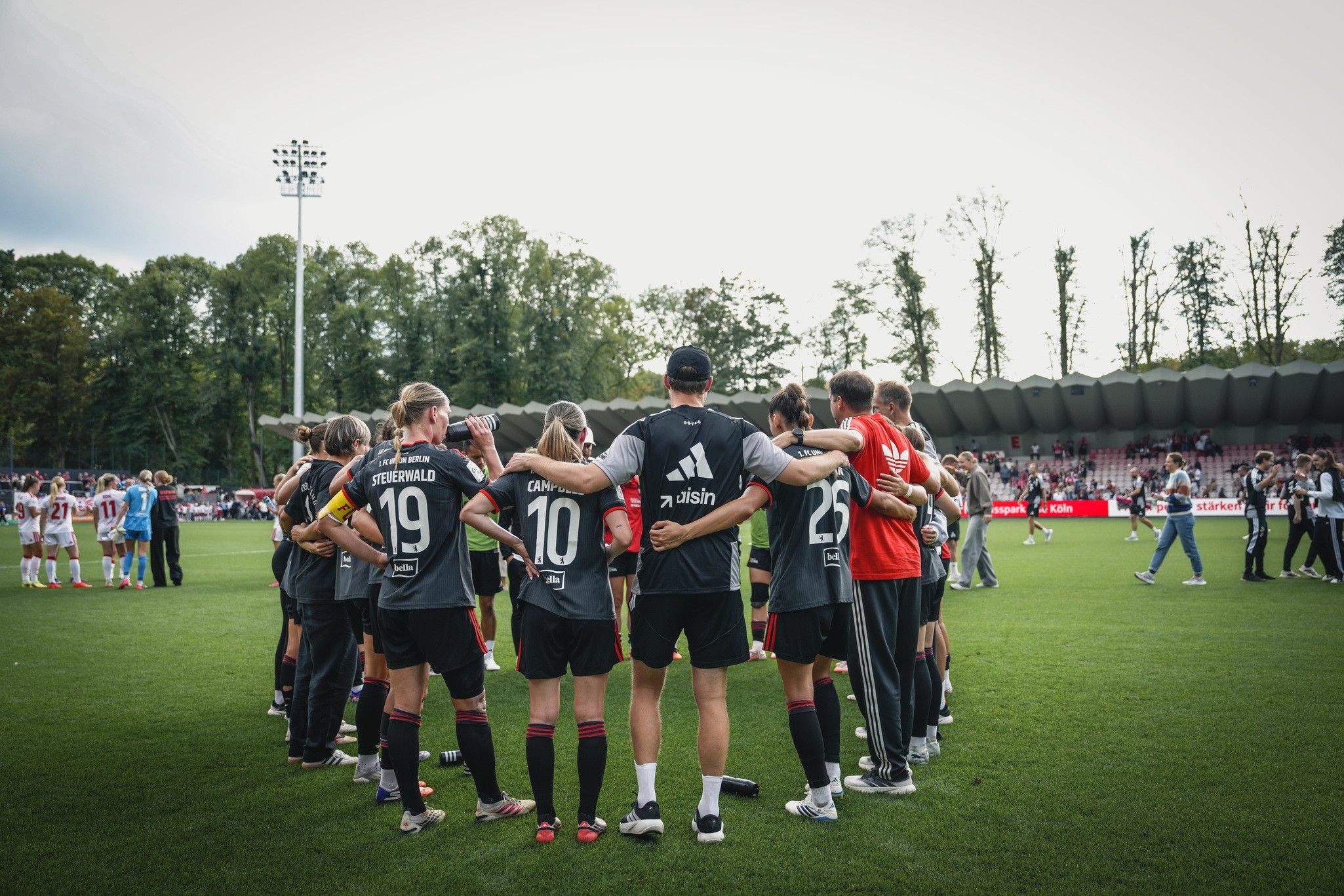 Spielerinnen einer Fußballmannschaft stehen einander umarmt auf dem Spielfeld, umgeben von einem Mannschaftstrainer und weiteren Teammitgliedern.