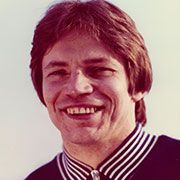 Ein lächelnder Mann mit braunen Haaren und einem gestreiften Kragen steht vor einem hellen Hintergrund.