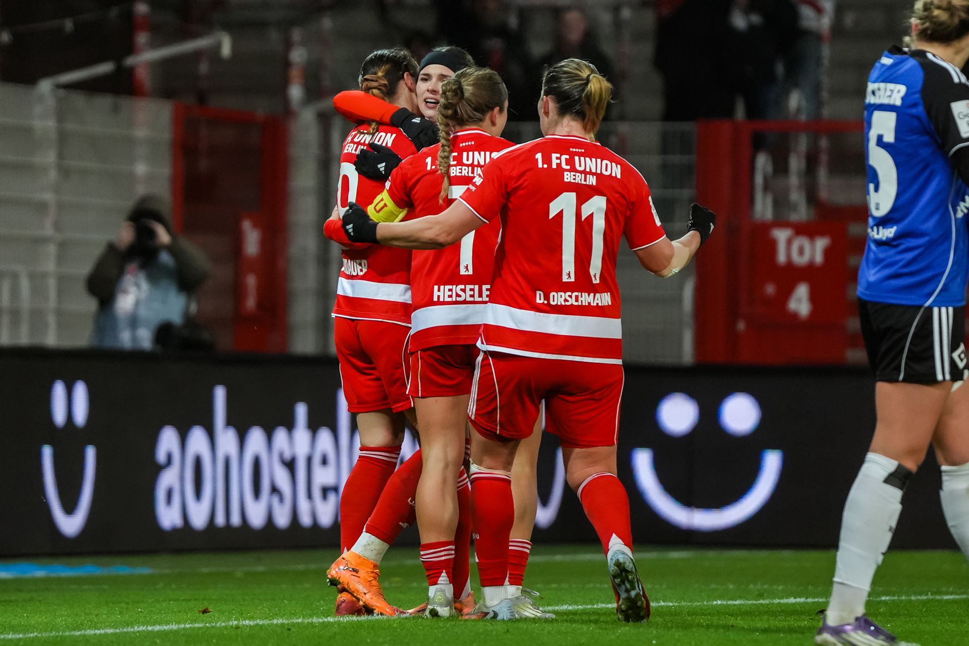 Drei Spielerinnen des 1. FC Union Berlin feiern einen Torerfolg und umarmen sich auf dem Spielfeld.