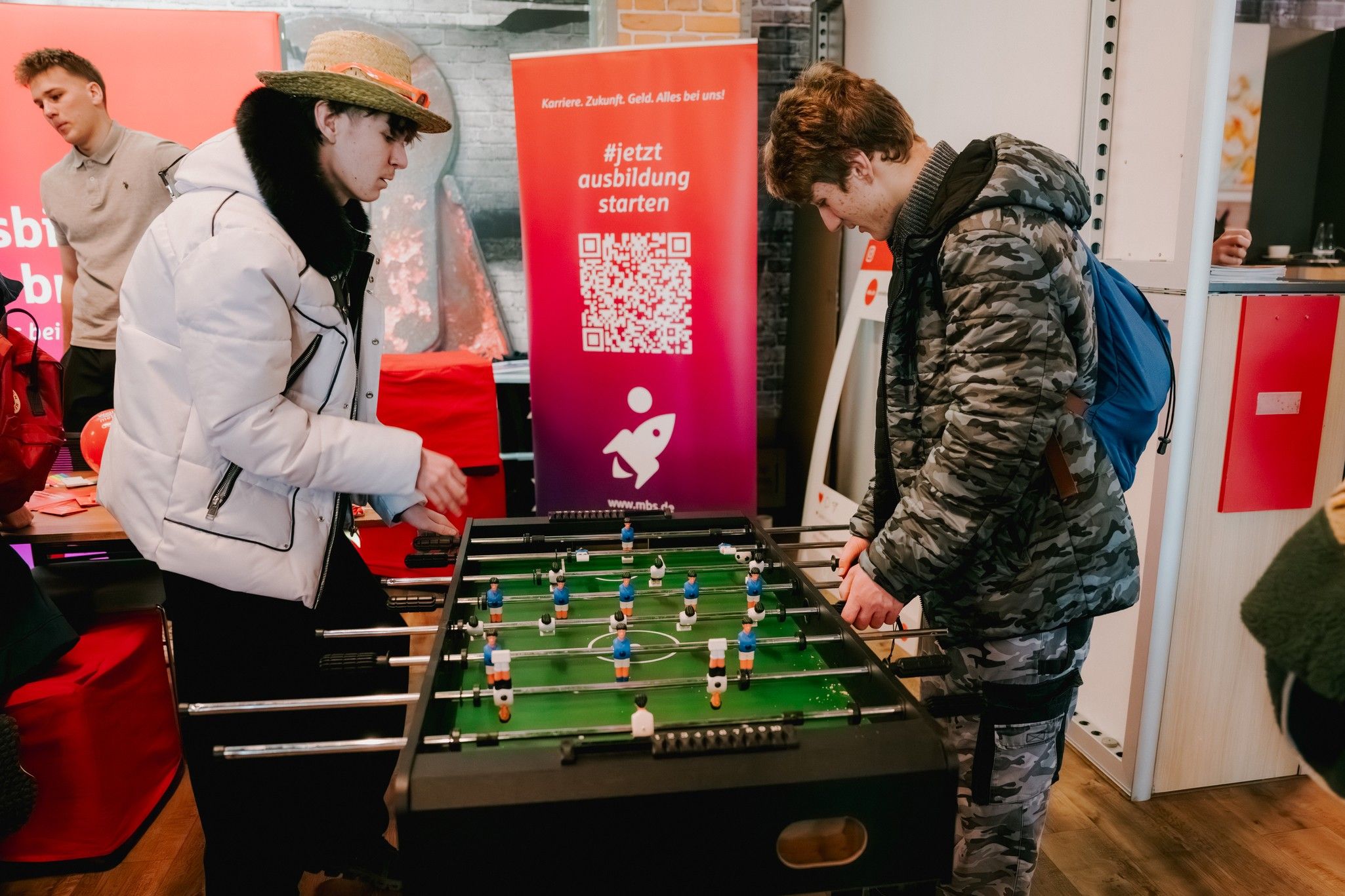 Zwei Personen spielen am Kickertisch in einem Ausstellungsraum mit roten Tischen im Hintergrund.
