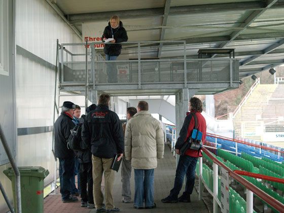 Eine Gruppe von fünf Personen steht in einem Stadion, während eine weitere Person auf einer oberen Plattform notiert.