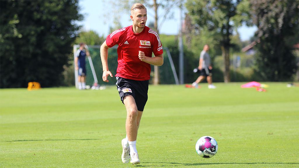 A football player in a red jersey is running with a ball on a training field in sunny weather.