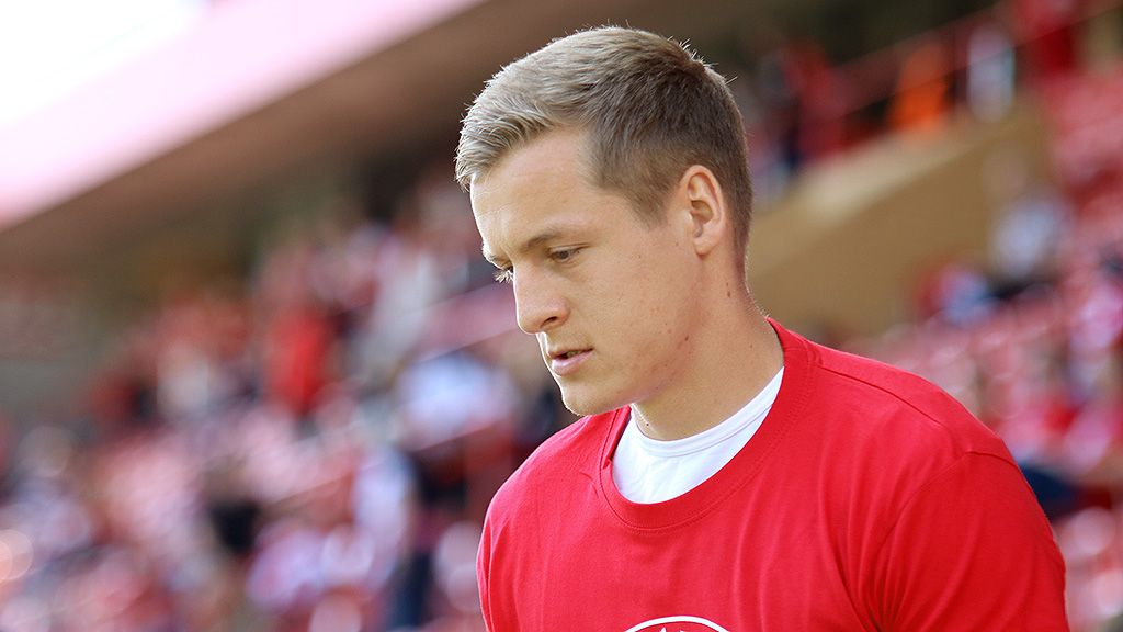 Ein junger Sportler trägt ein rotes T-Shirt und blickt nachdenklich, während er auf einem Stadionfeld steht.