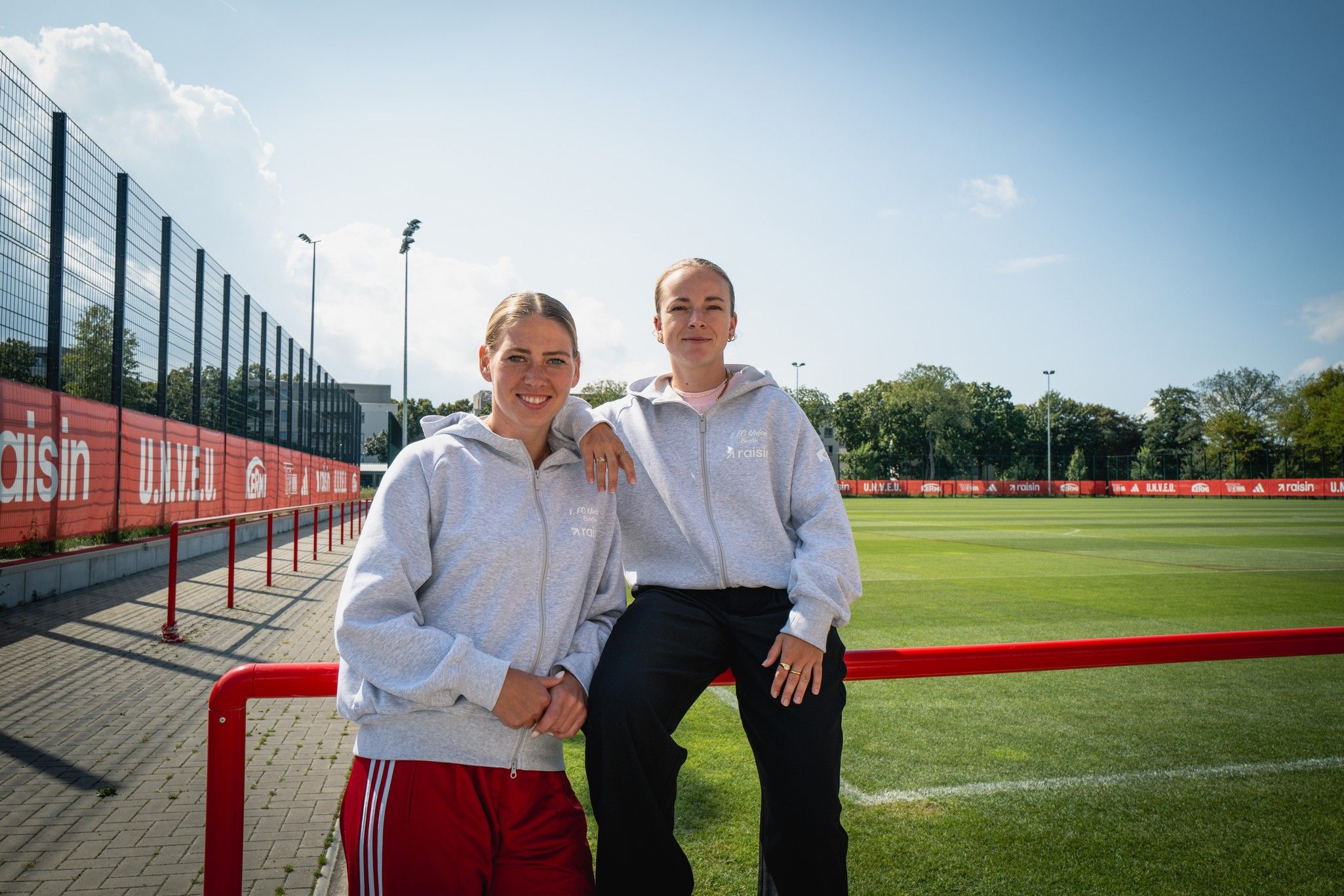 Zwei junge Frauen in grauen Sweatshirts stehen an einem Fußballplatz mit rotem Zaun und grünem Rasen, unter blauem Himmel.