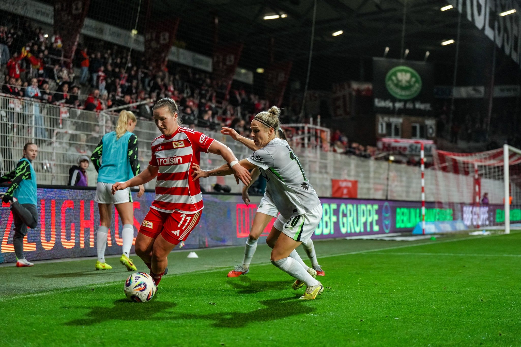 Eine Fußballspielerin im roten Trikot dribbelt den Ball, während eine gegnerische Spielerin im weißen Trikot sie verfolgt. Stadionatmosphäre im Hintergrund.
