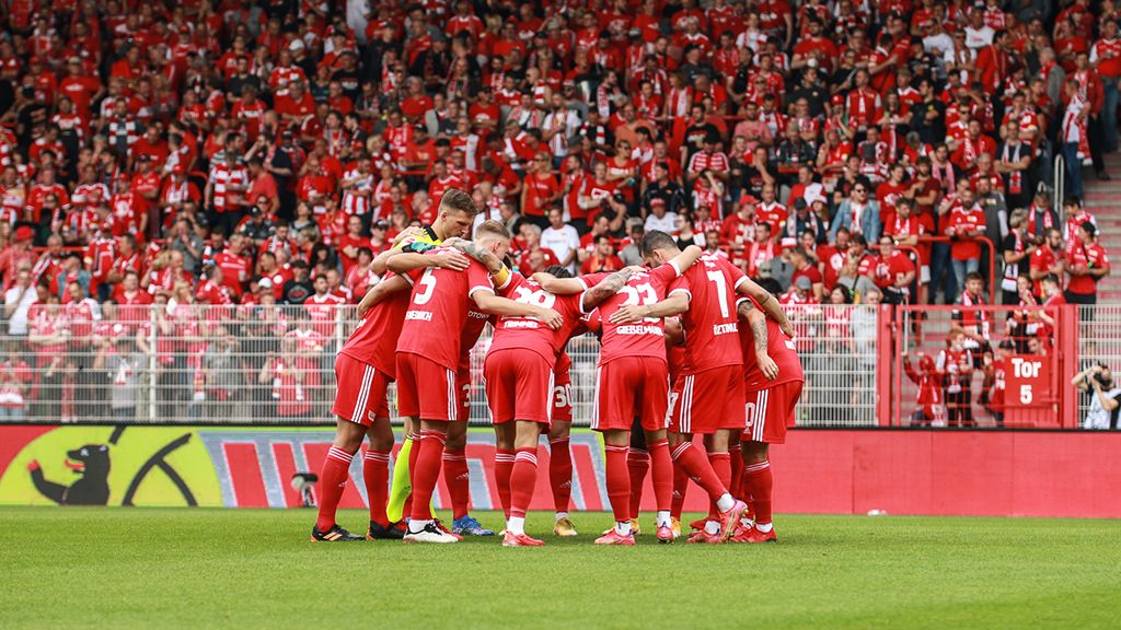 Mannschaft in roten Trikots bildet einen Kreis auf dem Spielfeld, Fans im Hintergrund unterstützen.