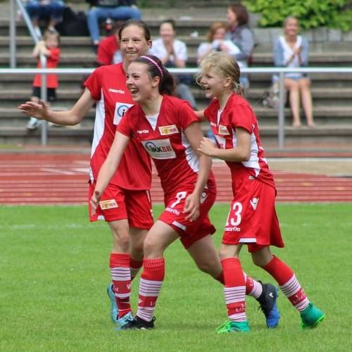 Drei junge Fußballspielerinnen in roten Trikots jubeln auf dem Spielfeld, während Zuschauer im Hintergrund zuschauen.