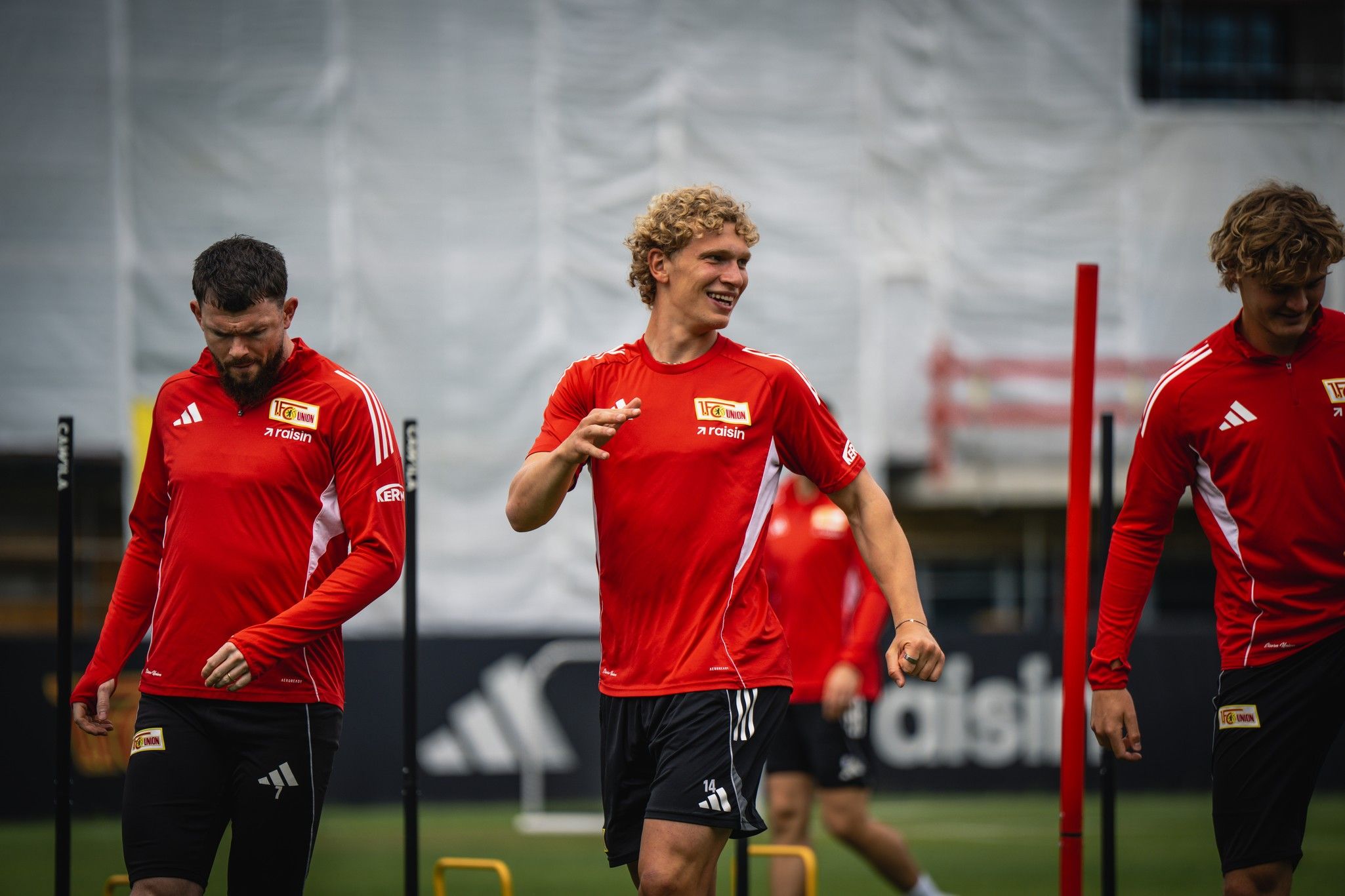 Drei Fußballspieler in roten Trikots trainieren auf einem Rasenplatz, mit Trainingsgeräten im Hintergrund.